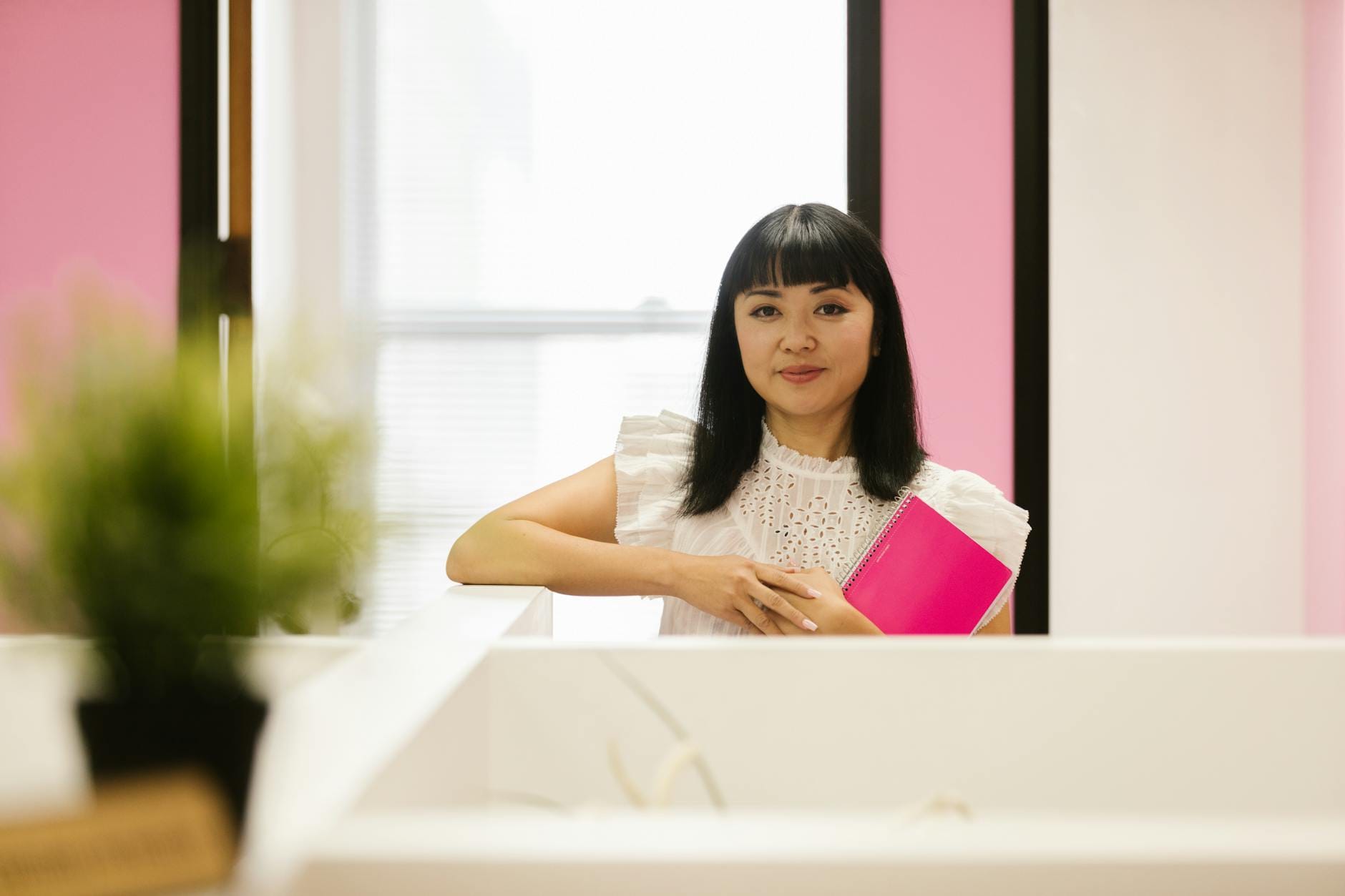 Asian woman in white top holding a pink notebook, smiling in a modern office setting.