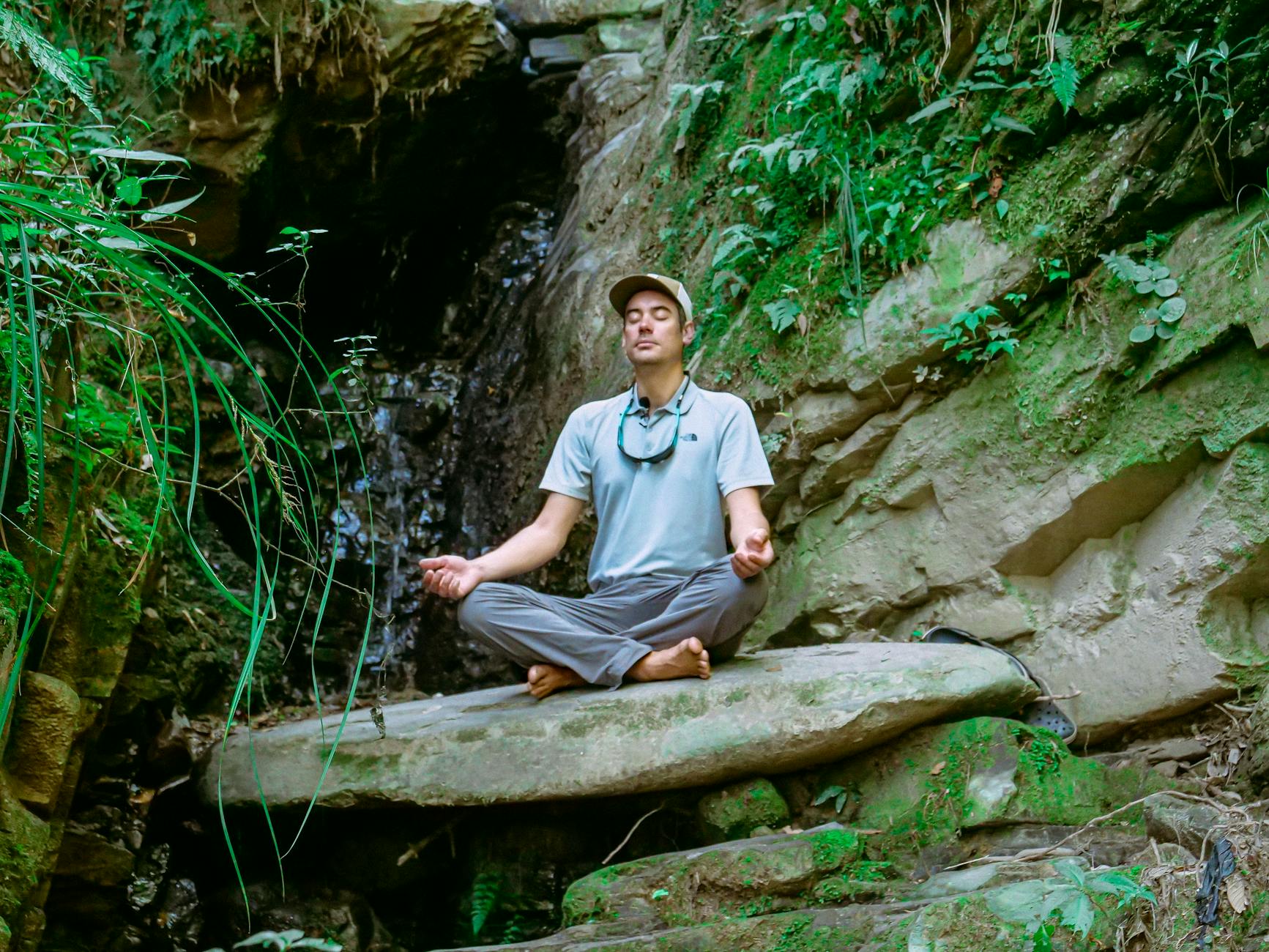 A man sits meditating on a rock by a waterfall in a serene natural setting, surrounded by lush greenery.
